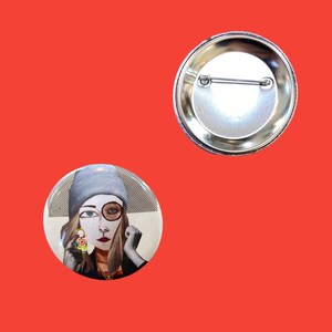 Peut inclure: Un bouton rond avec une femme portant un bonnet gris et un masque blanc avec une loupe sur un œil. La femme tient une petite poupée avec un motif floral.