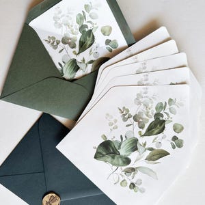 Pode incluir: Conjunto de papelaria com ilustrações botânicas. O conjunto inclui vários cartões brancos com desenhos de eucalipto verde e cinza, e envelopes verde escuro. Um envelope está aberto, revelando o interior do cartão. Um envelope azul escuro é selado com um selo de cera dourada.