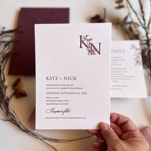 May include: A white wedding invitation with a burgundy monogram of the letters "K" and "N" intertwined with floral accents. The text reads "Kate + Nick" and includes the date "Saturday, September 23, 2023" and the time "Four O'Clock". The invitation also includes the text "Reception to follow" and "RSVP by August 10th at".