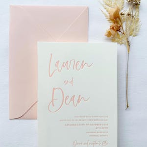 May include: A white wedding invitation with pink lettering. The invitation reads "Lauren and Dean" and includes the wedding date, time, and location. The text "Dinner and reception to follow" is also included.