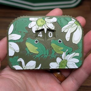 May include: A green and white decorative counter with two green frogs, white daisies, and the number 40. The counter is held in a hand and has the text "STUFF" in the background.