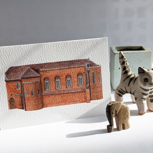 May include: A watercolour painting of a red brick building with a brown roof. The building has a series of windows and a door. The painting is on a white card.