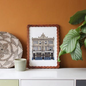 May include: A framed print of a detailed building facade, set in a brown, scalloped frame. The artwork depicts a multi-windowed building with a detailed roofline. The print is displayed on a white surface with a woven basket and a small green pot.