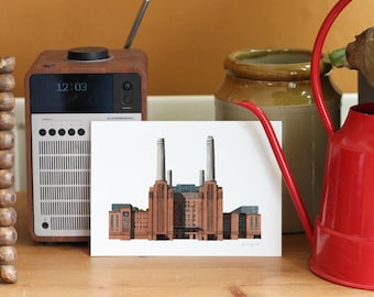May include: A print depicting Battersea Power Station, a landmark in London, with its distinctive chimneys. The artwork is on a white card. A wooden radio and a red watering can are also visible, creating a vintage aesthetic.