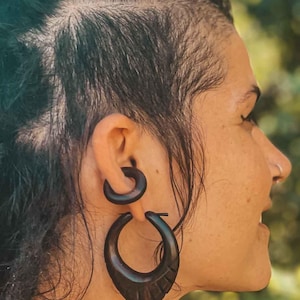 May include: A person with dark hair and a shaved side wearing large wooden hoop earrings. The earrings are dark brown and have a unique carved design.
