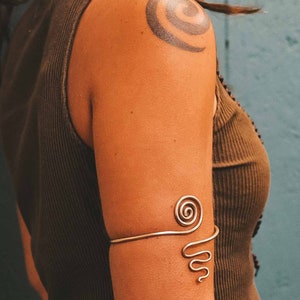 Op de afbeelding: Een goudkleurige armmanchet met een spiraalvormig ontwerp aan de bovenkant en een golvend lijnenontwerp eronder. De manchet wordt op de bovenarm gedragen. De persoon draagt een bruin tanktop. Een tatoeage is zichtbaar op de schouder.