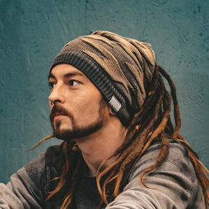 May include: A man with long dreadlocks wears a brown and gray beanie and a gray sweater. He has a tattoo on his arm and is sitting with his legs crossed.