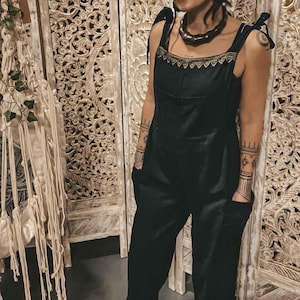 May include: A black jumpsuit with a brown beaded necklace and a brown turban. The jumpsuit has thin straps and a square neckline. The person is standing in front of a white wooden screen with intricate carvings.