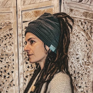 May include: A woman with long brown dreadlocks wears a dark gray knit beanie with a light blue tag. She is standing in front of a carved wooden screen.
