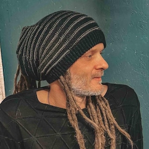May include: A person with long dreadlocks is wearing a black and grey striped beanie. The beanie is folded over at the top and the person is looking to the side.