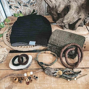 May include: A collection of handmade accessories, including a black knit beanie, a green scarf, brown bracelets, and a necklace with feathers and beads. The items are arranged on a wooden surface, showcasing a rustic aesthetic.
