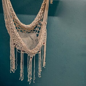 May include: A white macrame hammock with fringe hanging against a teal wall. The hammock has wooden beads woven into the design.