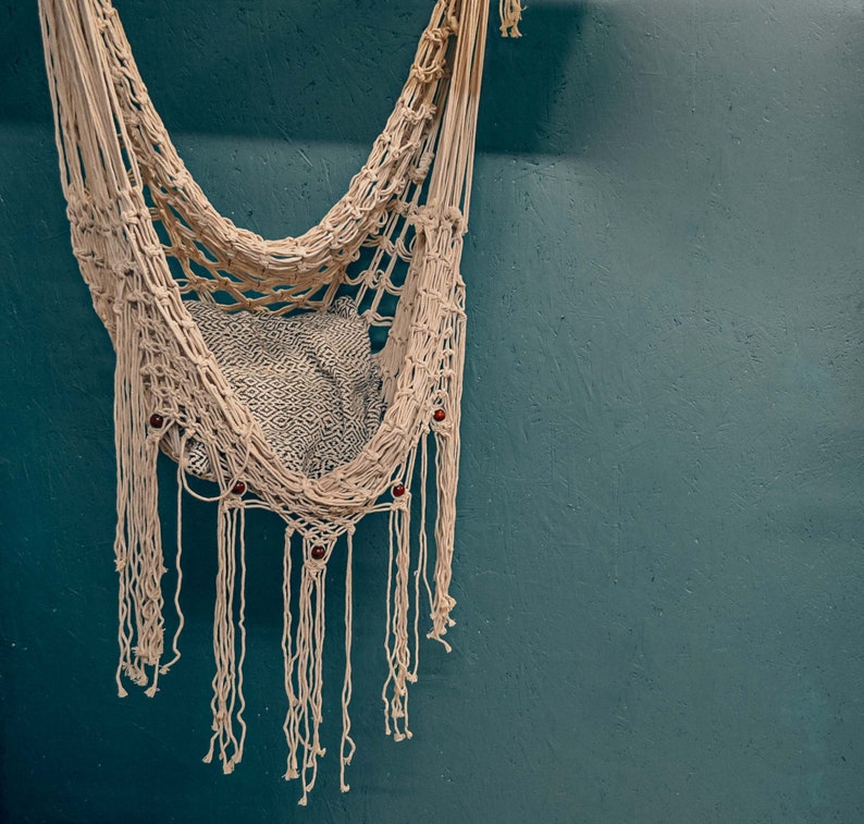May include: A white macrame hammock with fringe hanging against a teal wall. The hammock has wooden beads woven into the design.