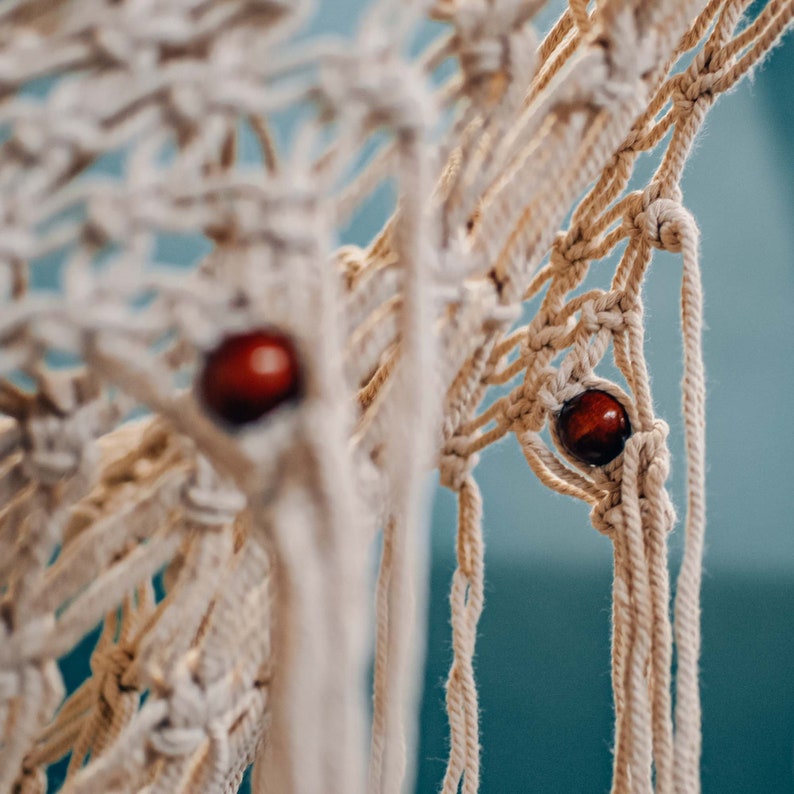 May include: A macrame wall hanging with a natural beige color and two red wooden beads. The macrame is made with a knotting technique and has a fringe at the bottom.