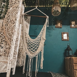May include: A white macrame hammock hanging from a wooden beam against a teal wall. The hammock is made of a thick, knotted rope and has a fringe at the bottom. The wall is decorated with carved wooden panels and mirrors.