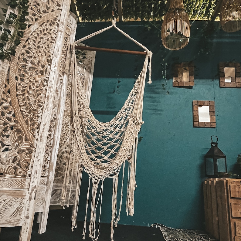 May include: A white macrame hammock hanging from a wooden beam against a teal wall. The hammock is made of a thick, knotted rope and has a fringe at the bottom. The wall is decorated with carved wooden panels and mirrors.