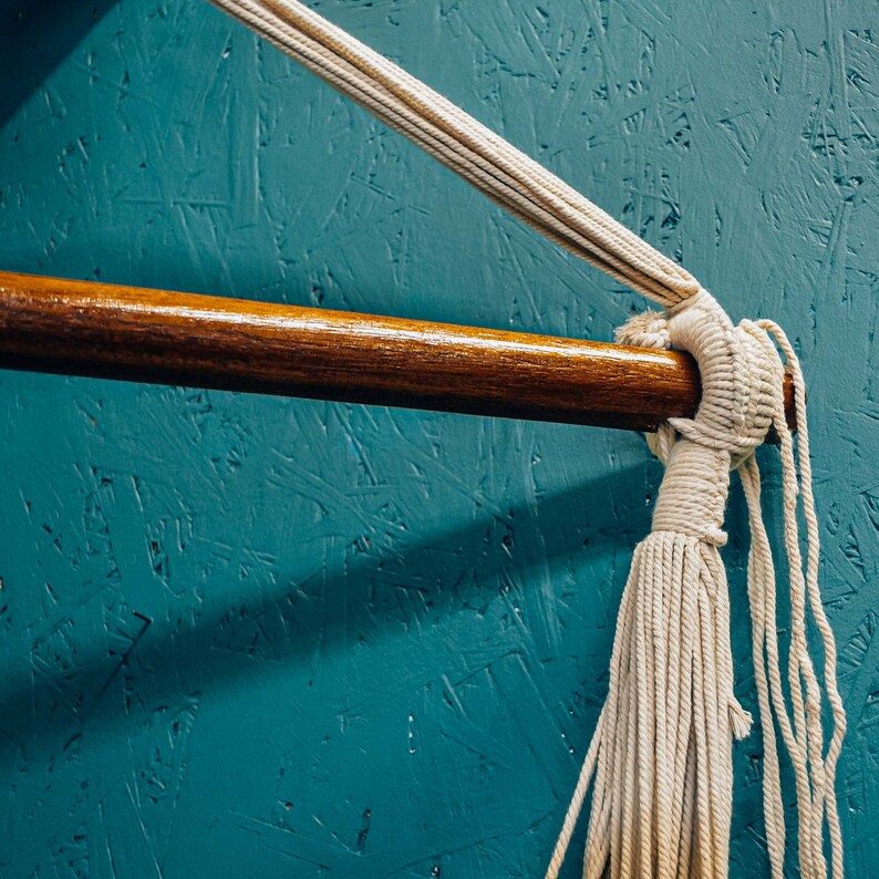 May include: A white cotton rope hammock is tied to a wooden bar against a teal blue wall. The rope is knotted and has long tassels.