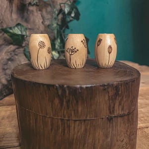 Könnte beinhalten: Drei Holzperlen mit floralen Designs. Die Perlen sind hellbraun und haben eine natürliche Holzoptik. Die Designs sind schlicht und elegant und zeigen zarte Blumen und Blätter.