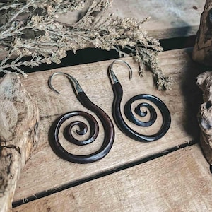 Puede incluir: Un par de pendientes en espiral de color marrón oscuro con ganchos plateados. Los pendientes están hechos de madera y tienen un acabado liso y pulido. El diseño en espiral es una forma clásica y elegante.