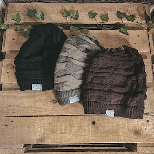May include: Three knit beanies in black, gray, and dark brown, each featuring a cable knit pattern and a ribbed cuff. A small rectangular white label with the text "Crafter" in blue script is attached to the bottom edge of each beanie.