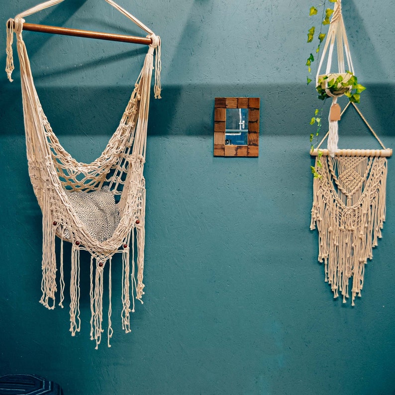 May include: Two macrame wall hangings, one is a hammock style chair and the other is a traditional wall hanging. Both are white and have a natural wood accent. The wall is a teal blue color.