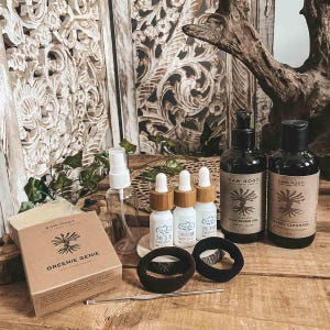 May include: A selection of Raw Root skincare items, including bottles, droppers, a soap bar, and hair ties. The products are displayed on a wooden surface with decorative wooden panels behind. The soap box reads "Greenie Genie."