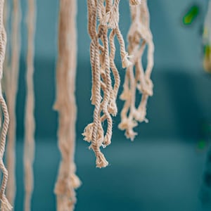 May include: Close-up of a macrame wall hanging with natural colored cotton cords. The cords are knotted and frayed at the ends, creating a textured and bohemian look.