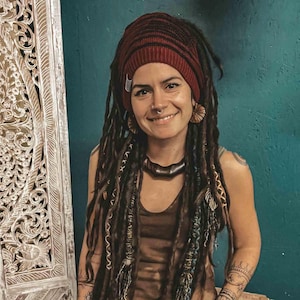 May include: A woman with long brown dreadlocks wears a maroon beanie and a brown tank top. She has a nose ring and wooden earrings.