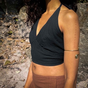 May include: A black halter-style crop top with a V-neckline and a wrap-front design, paired with a gold-toned metal arm cuff featuring spiral ends. The top is sleeveless and cropped at the midriff, while the arm cuff sits above the elbow.
