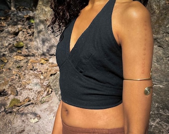 May include: A black halter-style crop top with a V-neckline and a wrap-front design, paired with a gold-toned metal arm cuff featuring spiral ends. The top is sleeveless and cropped at the midriff, while the arm cuff sits above the elbow.