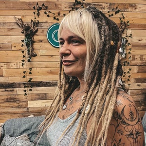 May include: A woman with long blonde hair and dreadlocks, wearing a grey tank top and a necklace. She has a tattoo on her right shoulder and arm, featuring a crescent moon and floral designs.