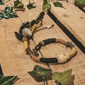 Peut inclure: Un bracelet tressé vert, marron et jaune avec une perle en bois et un charme en métal. Le bracelet est sur une surface en bois avec des feuilles vertes.