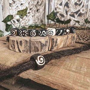 May include: A collection of black and white hair beads with various spiral and geometric designs. The beads are displayed on a wooden surface, with one bead attached to a dreadlock. The beads are approximately 0.75 inches in diameter.