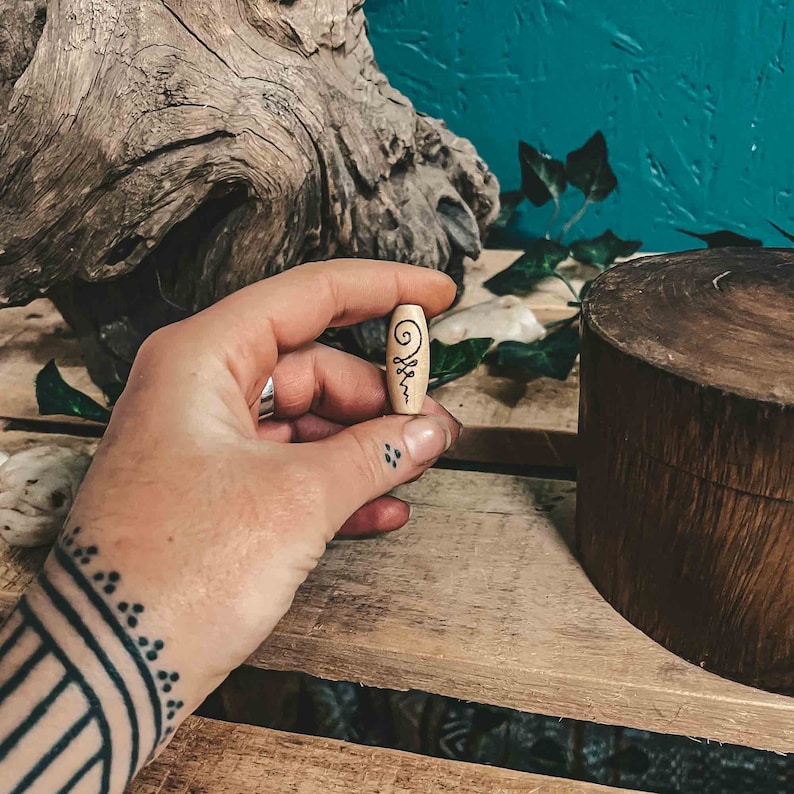 May include: A small wooden bead with a black and white symbol carved into it. The bead is being held by a hand with a tattoo.