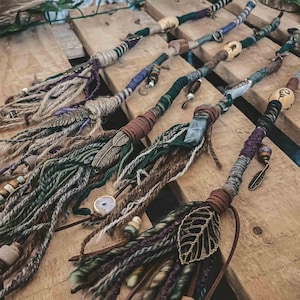 Peut inclure: Plusieurs extensions de dreadlocks faites à la main. Chaque extension est ornée d'éléments en bois ou en métal, avec des couleurs vertes, marron, violettes et crème. Des breloques en forme de plumes et de feuilles sont également présentes. Les extensions sont disposées sur une surface en bois.