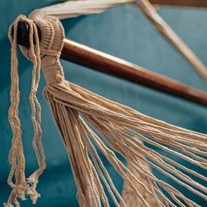 May include: Close-up of a beige hammock made of woven rope, hanging from a wooden bar against a teal blue wall.