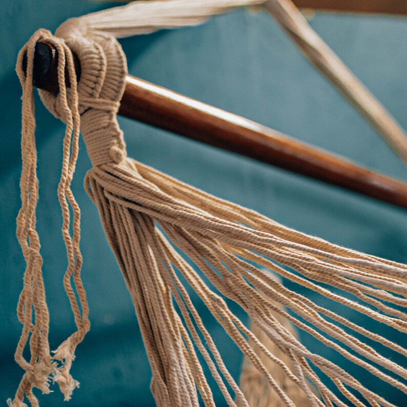 May include: Close-up of a beige hammock made of woven rope, hanging from a wooden bar against a teal blue wall.