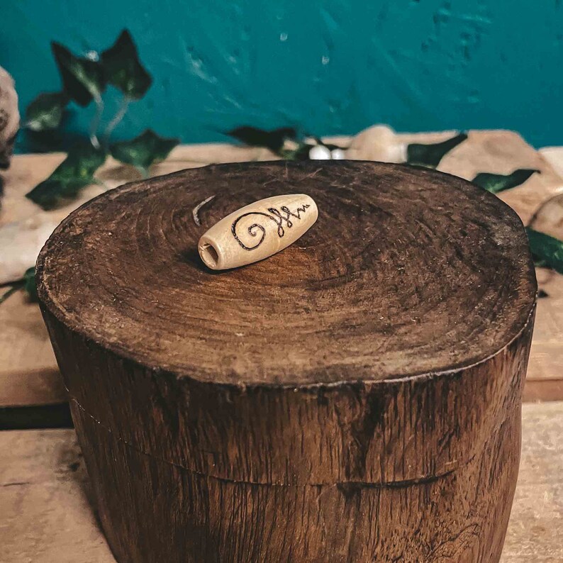 May include: A small, wooden bead with a carved spiral design. The bead is light brown in color and is sitting on a wooden surface.