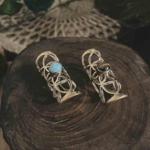 May include: Two ornate, gold-colored finger rings. One ring features a light blue gemstone, the other a brown gemstone. The rings have an open, geometric design. The rings are displayed on a dark, wooden surface.