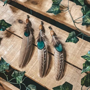 Op de afbeelding: Drie bruine veren met turquoise accenten en houten kralen aan de bovenkant. De veren zijn horizontaal op een houten oppervlak gerangschikt.