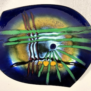 May include: A decorative, abstract ceramic plate with a dark blue border. The center features a design with green, yellow, and red accents. The plate has a unique, irregular shape and a glossy finish.