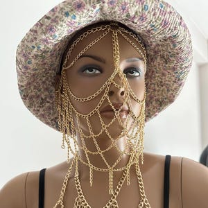 Puede incluir: Un arnés de cuerpo de cadena dorada con una máscara facial a juego de cadena dorada. La máscara facial es un diseño delicado de cadena que cubre la frente, los ojos, la nariz y la boca. El arnés de cuerpo está hecho de múltiples capas de cadena que caen sobre los hombros y el pecho.