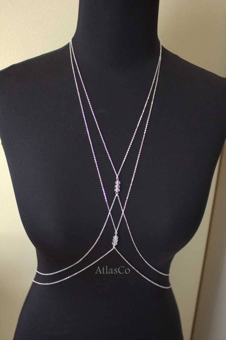 Op de afbeelding: Een zilveren body chain met drie strengen en kristallen accenten. De ketting is gelaagd en kruist elkaar aan de voorkant. De ketting is op een zwart manikin.