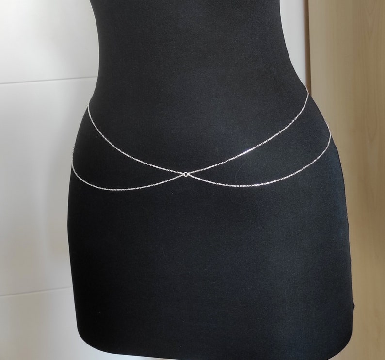 May include: A silver body chain with two strands that cross in the front. The chain is worn on a black mannequin.