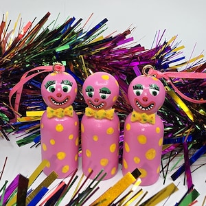 May include: Three pink wooden clown ornaments with yellow polka dots and bow ties. Each ornament has a painted face with green eyes and a smiling mouth. Pink ribbon loops are attached for hanging.
