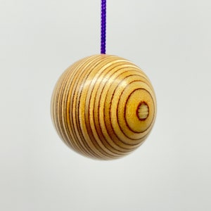 May include: A wooden ball with a striped pattern, hanging from a purple string. The ball is light brown with darker brown lines. The object is likely a toy or decorative item. The ball's diameter is approximately 5 cm.