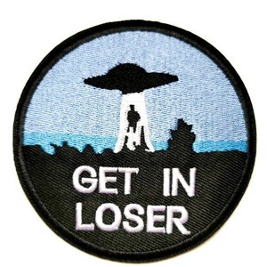 May include: A black and blue embroidered patch with a UFO hovering over a person standing on a hill. The text "GET IN LOSER" is written in white below the UFO.