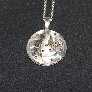 May include: Silver pendant necklace featuring a circular watch mechanism. The pendant showcases intricate gears, ruby-colored jewels, and the inscription "ROGER WATCH CO." on a silver chain. The background is a dark gray.