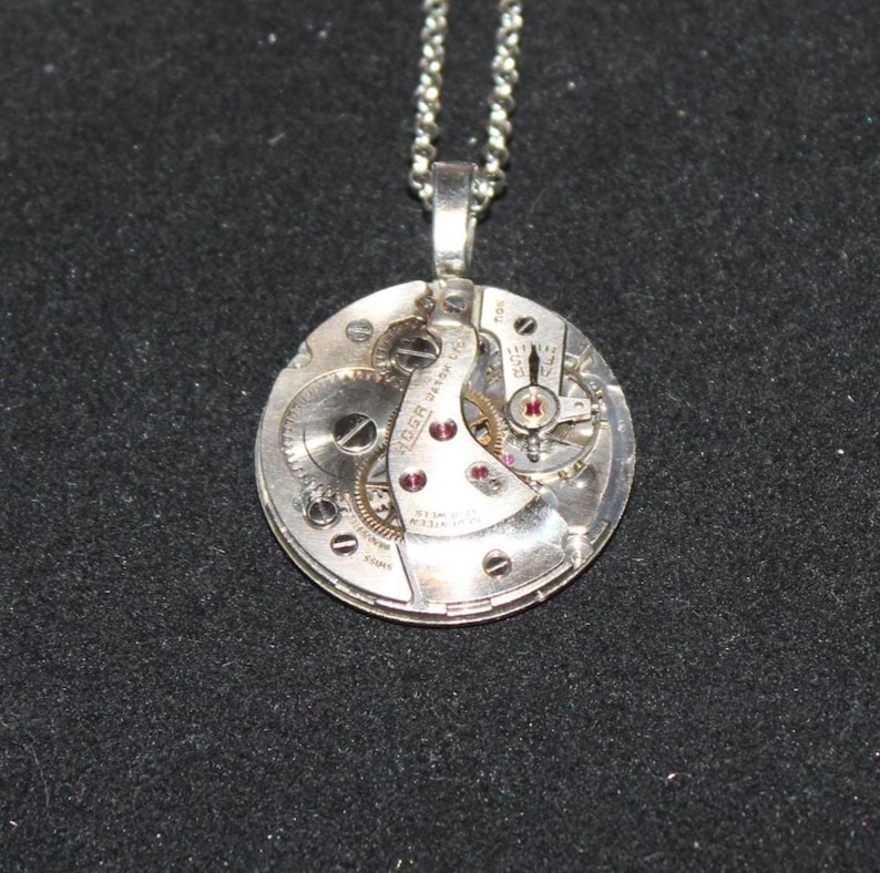 May include: Silver pendant necklace featuring a circular watch mechanism. The pendant showcases intricate gears, ruby-colored jewels, and the inscription "ROGER WATCH CO." on a silver chain. The background is a dark gray.
