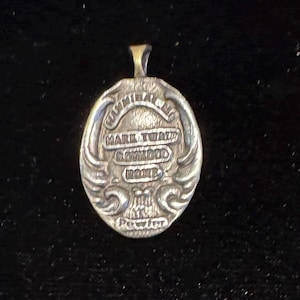 May include: Oval-shaped silver pendant with the text "MARK TWAIN BOYHOOD HOME" and "Pewter" at the bottom. The pendant has a loop at the top for a chain and decorative scrollwork around the edges. The pendant is on a black background.